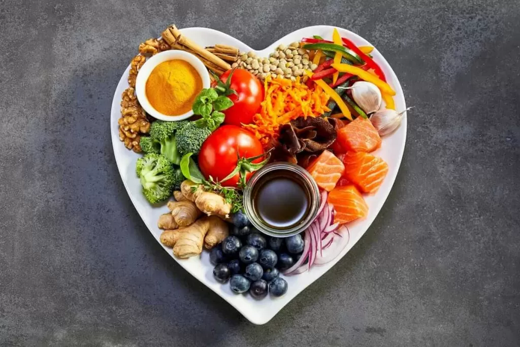 تصویری از یک ظرف غذای سالم که ظرف به شکل قلب سفید می باشد و در آن انواع میوه جات و سبزی جات سالم از جمله پرتقال، گوجه فرنگی، ترب، دانه های انگور، هویچ، کلم بروکلی، زنجویل، دارچین، زرچوبه، گردو، سیر و ... می باشد. این تصویر نماد رژیم غذایی سالم مناسب برای کاهش اضطراب و استرس می باشد.
